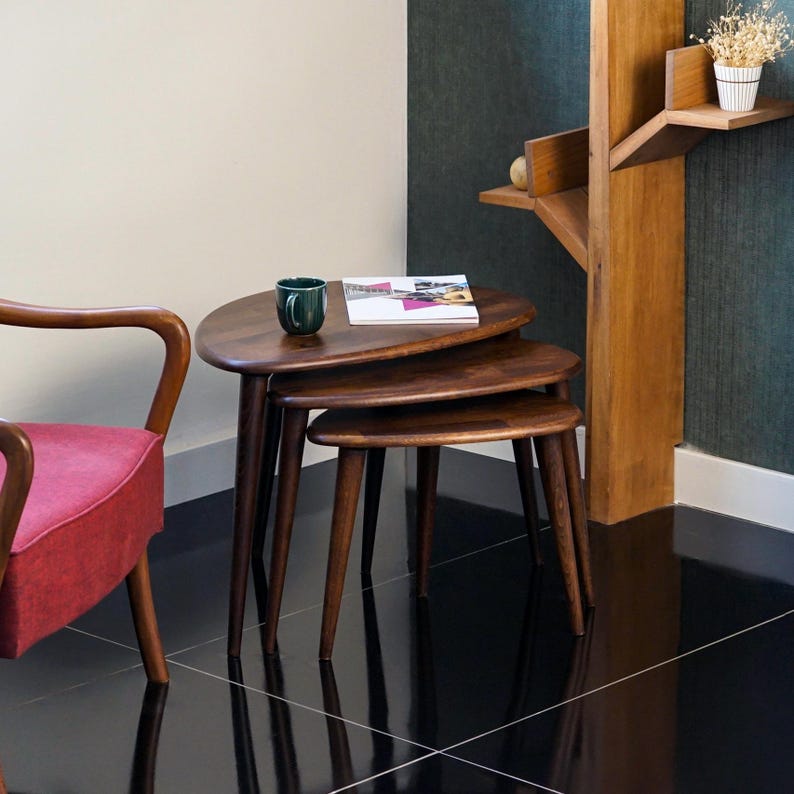 May include: Set of three nesting tables in a warm brown finish. The tables have a rounded, organic shape and tapered legs. A dark green mug and a magazine rest on the top table. The tables are a stylish addition to any living space.