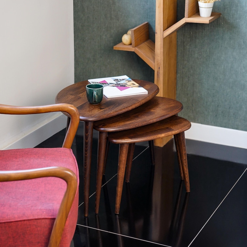 May include: A set of three wooden nesting tables with oval tops and tapered legs. The tables are a rich brown color and are arranged in a staggered formation. A green cup and a magazine rest on the largest table. A red chair is in the foreground.
