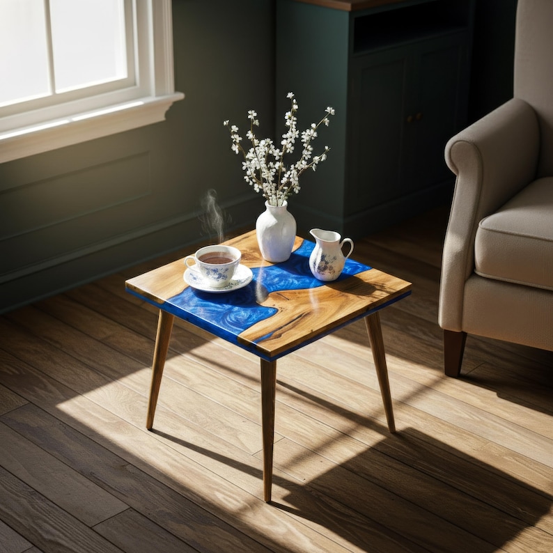 May include: A square wooden side table with a blue epoxy resin design. The table has four wooden legs and a wooden top with a blue resin river. A cup of tea, a vase of flowers, and a creamer are on the table.