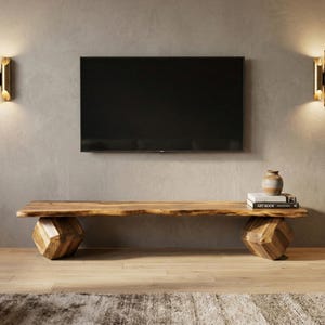 May include: A natural wood TV console with a live edge top and geometric cube-shaped legs. A black flat-screen TV is mounted on the gray wall above. Decorative sconces flank the TV. A vase and books sit on the console.