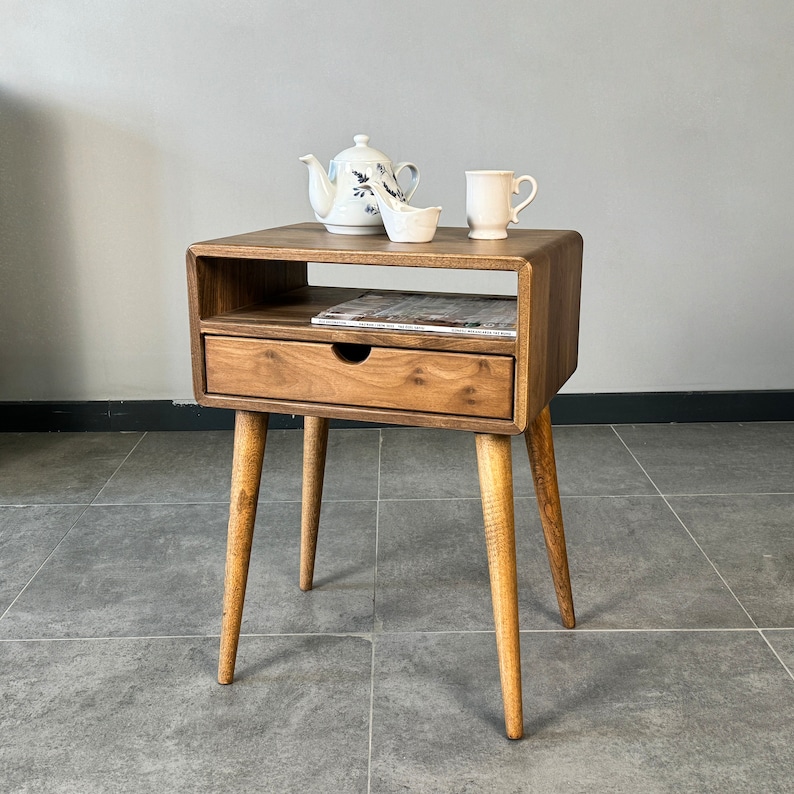 Op de afbeelding: Een houten bijzettafel met een lade en een plank. De tafel heeft een lichtbruine afwerking en vier taps toelopende poten. Een witte theepot, een witte mok en een witte schotel staan op de bovenste plank.