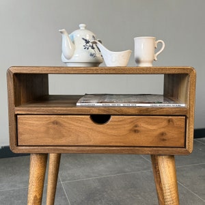Op de afbeelding: Een houten bijzettafel met een lade en een plank. De tafel heeft een lichtbruine afwerking en is versierd met een witte theepot, een witte roomkan en een witte mok. Een tijdschrift ligt op de plank. De tijdschriftcover luidt "ELLE DECORATION HAZİRAN / EYL&Uuml;L 2023 YAZ &Ouml;ZEL SAYISI G&Uuml;NEŞLİ MEKANLARDA YAZ R&Uuml;YASI"