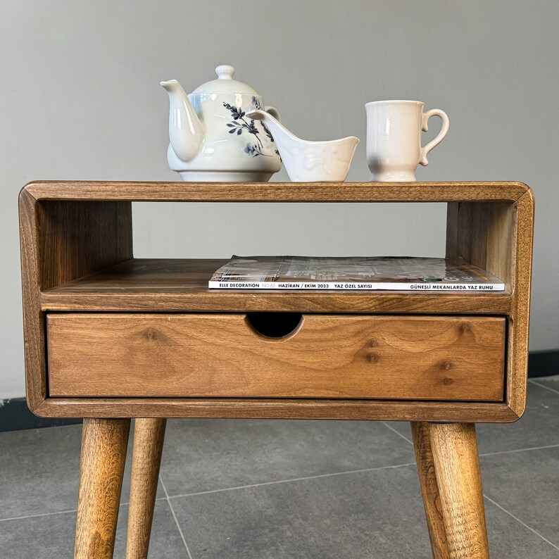 Op de afbeelding: Een houten bijzettafel met een lade en een plank. De tafel heeft een lichtbruine afwerking en is versierd met een witte theepot, een witte roomkan en een witte mok. Een tijdschrift ligt op de plank. De tijdschriftcover luidt "ELLE DECORATION HAZİRAN / EYL&Uuml;L 2023 YAZ &Ouml;ZEL SAYISI G&Uuml;NEŞLİ MEKANLARDA YAZ R&Uuml;YASI"