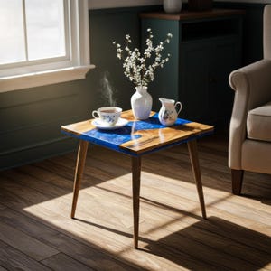 May include: A small, square side table with a wooden top inlaid with blue resin. The table has four angled wooden legs. A cup of tea, a creamer, and a vase of white flowers are on the table.
