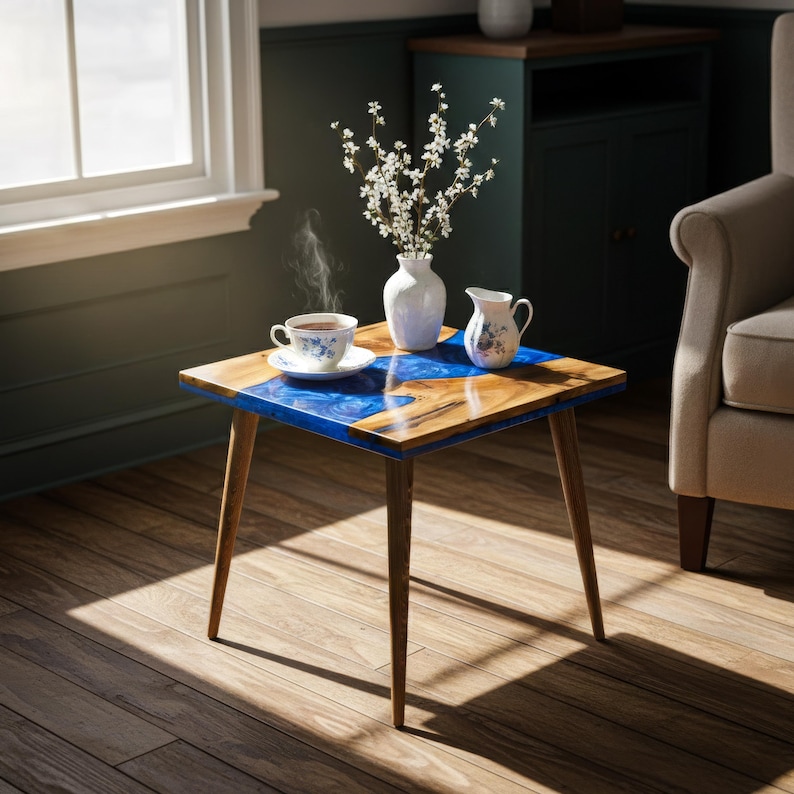 May include: A small, square side table with a wooden top inlaid with blue resin. The table has four angled wooden legs. A cup of tea, a creamer, and a vase of white flowers are on the table.