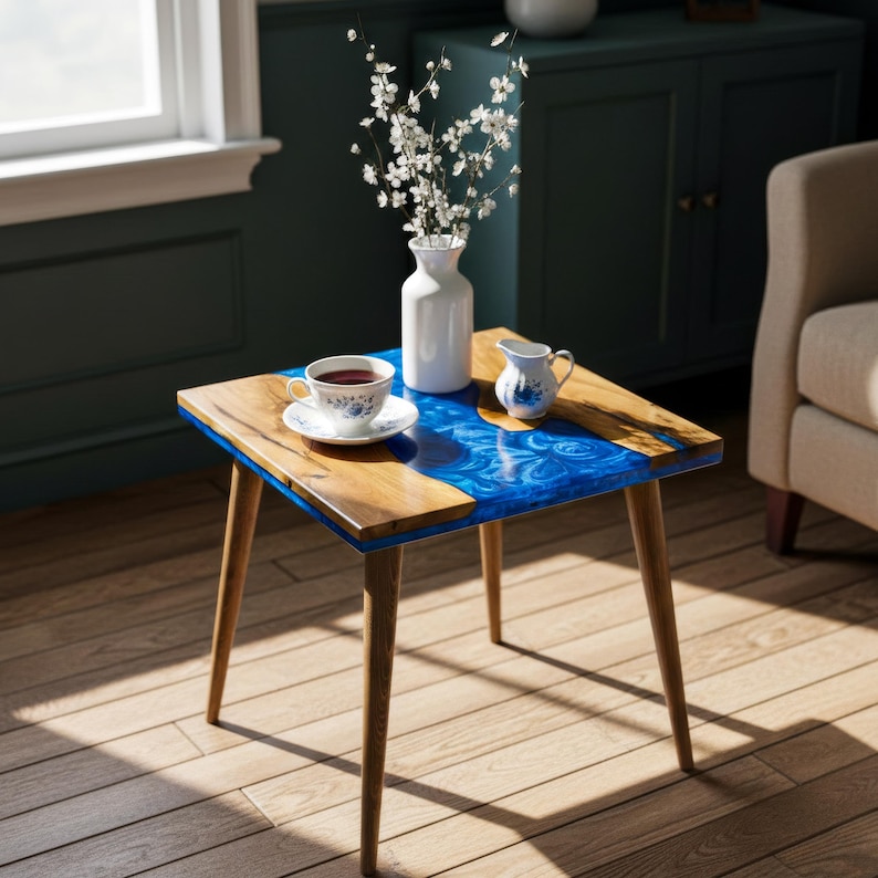 May include: A small, square side table with a wooden frame and legs. The tabletop features a combination of natural wood and a vibrant blue resin design. A white vase with flowers, a teacup, and a creamer sit on the table.