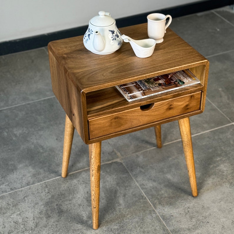 Op de afbeelding: Een houten bijzettafel met een lade en een plank. De tafel heeft vier poten en is gemaakt van donker hout. Er liggen een boek, een theepot, een kopje en een lepel op de tafel.