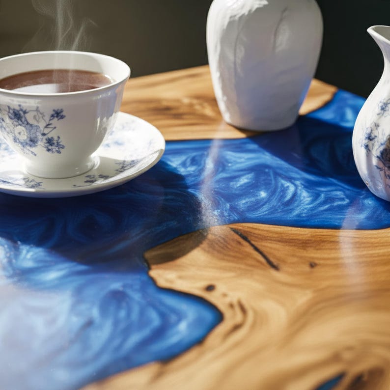May include: A close-up of a wooden table with a blue epoxy resin river. A white teacup and saucer with blue floral designs sits on the table, with steam rising from the cup. Two white ceramic vases are also on the table.