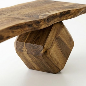 May include: A wooden bench featuring a thick, horizontal slab seat with a natural, uneven live edge. The seat is supported by a single, substantial base carved into a geometric, multi-faceted shape with visible wood grain patterns.