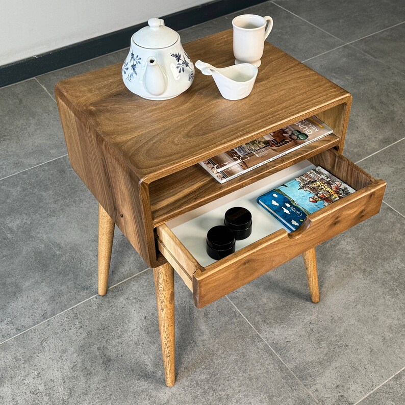 Op de afbeelding: Een houten bijzettafel met twee planken en een lade. De tafel heeft een lichtbruine afwerking en is versierd met een witte theepot, een witte mok en een tijdschrift. De lade is open en bevat een blauw boek en twee kleine ronde containers.