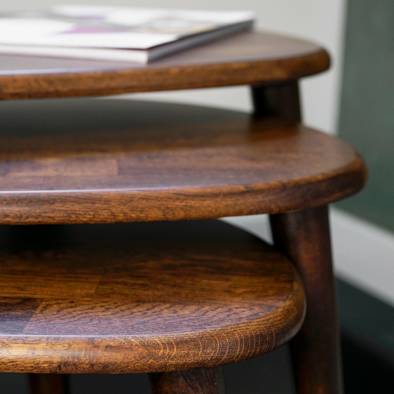 May include: A set of three nesting tables made of dark brown wood. The tables have a rounded triangular shape and are stacked, with a book resting on the top table. The wood grain is visible, adding texture to the furniture.