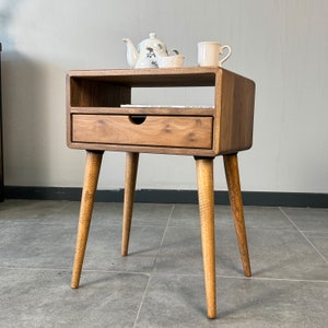 Op de afbeelding: Een houten bijzettafel met een lade en een plank. De tafel heeft een lichtbruine afwerking en vier taps toelopende poten. Een theepot, een kleine kom en een mok staan op de bovenste plank.