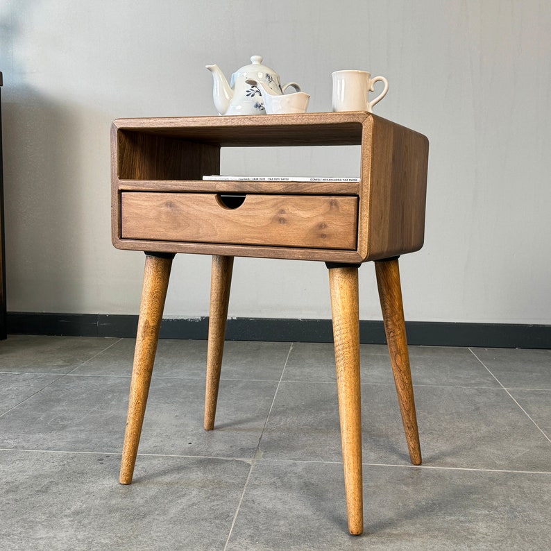 Op de afbeelding: Een houten bijzettafel met een lade en een plank. De tafel heeft een lichtbruine afwerking en vier taps toelopende poten. Een theepot, een kleine kom en een mok staan op de bovenste plank.