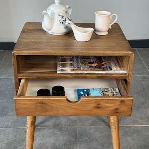 Op de afbeelding: Een houten bijzettafel met een lade en een plank. De tafel heeft een lichtbruine afwerking en afgeronde randen. De lade is open en bevat twee kleine potjes en een boek. De plank is leeg. De tafel staat op een grijze tegelvloer.