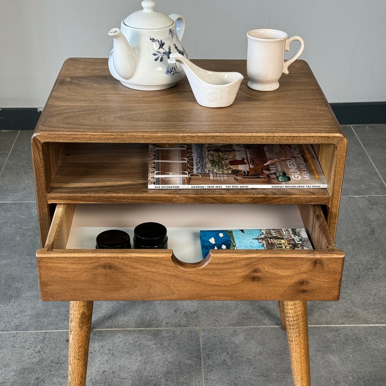 Op de afbeelding: Een houten bijzettafel met een lade en een plank. De tafel heeft een lichtbruine afwerking en afgeronde randen. De lade is open en bevat twee kleine potjes en een boek. De plank is leeg. De tafel staat op een grijze tegelvloer.