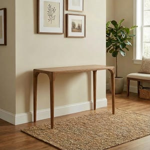 May include: A light brown wooden console table with four curved legs. The table is placed on a woven jute rug. The room has framed artwork on the wall and a potted plant.