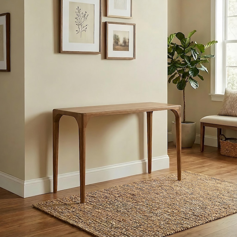 May include: A light brown wooden console table with four curved legs. The table is placed on a woven jute rug. The room has framed artwork on the wall and a potted plant.