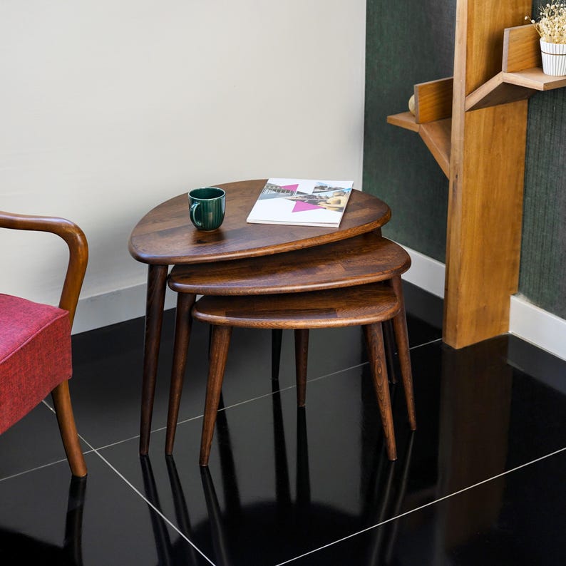 May include: Set of three nesting tables with a dark wood finish. The tables have a unique, rounded triangular shape and tapered legs. A green mug and a magazine rest on the top table. The tables are arranged on a black tiled floor.