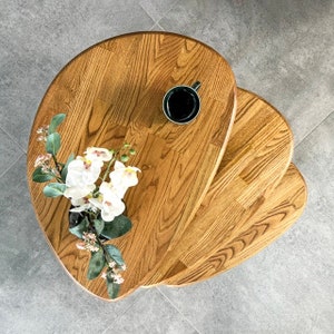 May include: A set of three wooden nesting tables with a natural wood finish. The tables are shaped like an oval and have a unique, organic design. The top table has a small, black cup on it.