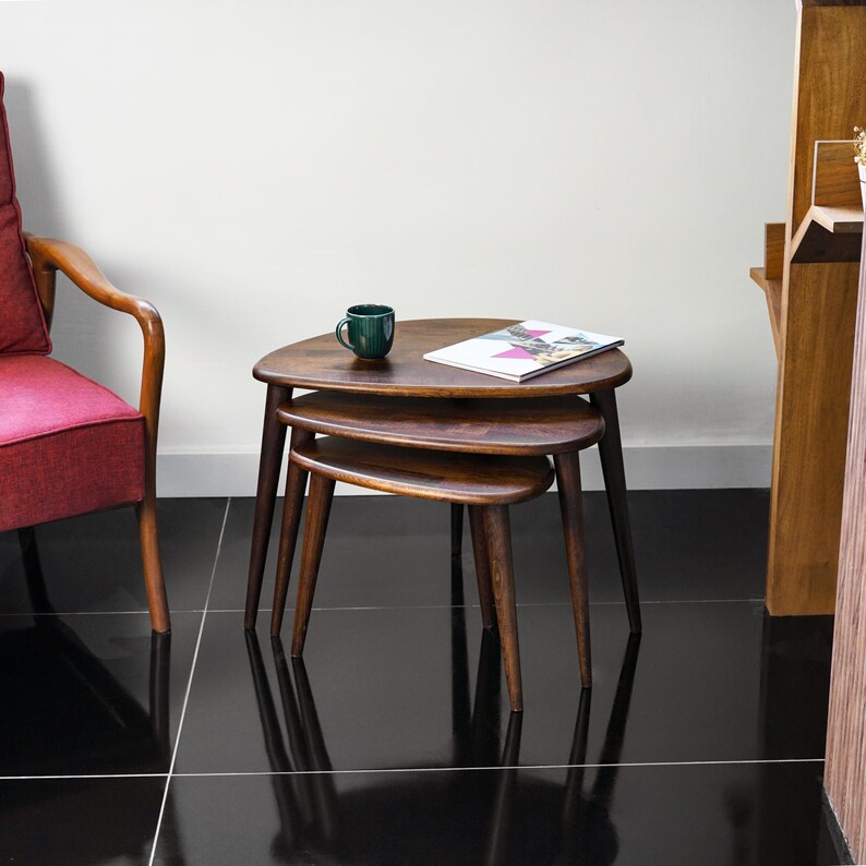 May include: A set of three wooden nesting tables with a dark brown finish. The tables have triangular tops and tapered legs. A green mug and a magazine are on the top table. The tables are arranged in a living room setting.