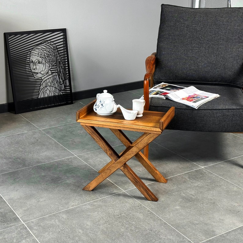 The wooden side table is placed beside an amchair. The side table is handmade and made by walnut wood. It is tray like shape and holding a teapot.