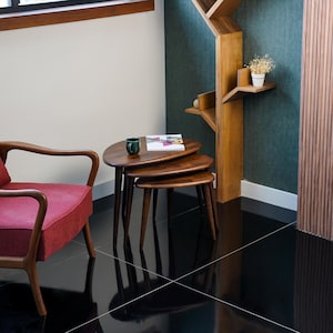 May include: A set of three wooden nesting tables with a dark finish. The tables have a rounded, organic shape and tapered legs. A red armchair and a wooden bookshelf are in the background. A green mug and a book are on top of the largest table.