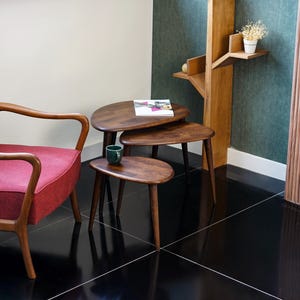 May include: A set of three wooden nesting tables with a dark finish. The tables have a rounded, organic shape and are supported by slender, tapered legs. A red armchair is visible on the left, and a green mug sits on the smallest table.