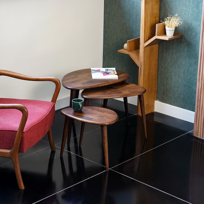 May include: A set of three wooden nesting tables with a dark finish. The tables have a rounded, organic shape and are supported by slender, tapered legs. A red armchair is visible on the left, and a green mug sits on the smallest table.