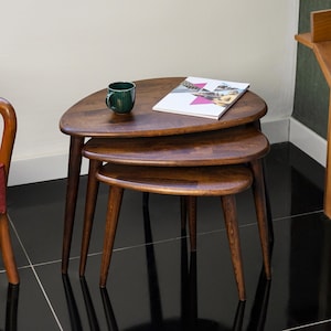Può includere: Un set di tre tavolini impilabili in legno con finitura marrone scuro. Ogni tavolo ha un piano triangolare e gambe affusolate. Una tazza verde e una rivista sono sul tavolo superiore. I tavoli sono impilati.