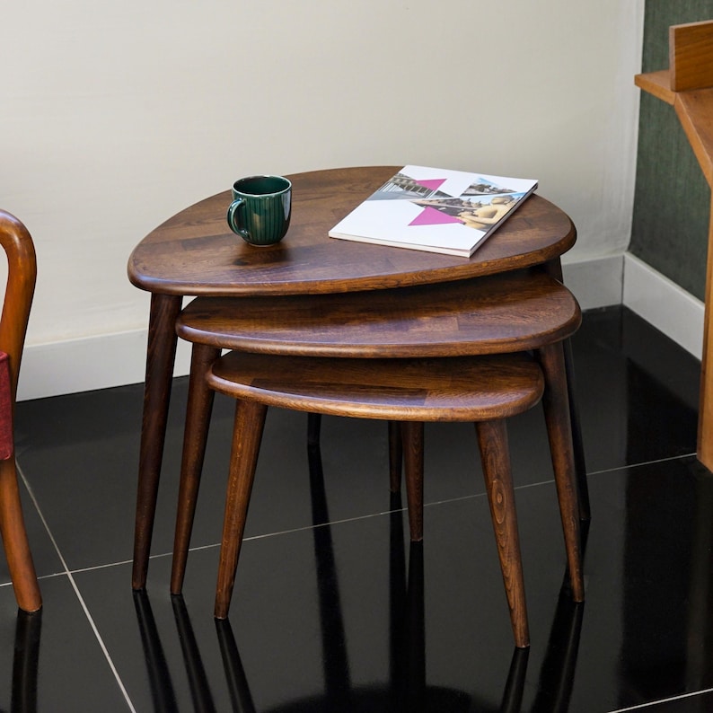 May include: A set of three wooden nesting tables with a dark brown finish. Each table has a triangular top and tapered legs. A green mug and a magazine are on the top table. The tables are arranged in a stacked configuration.