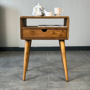 Op de afbeelding: Een houten bijzettafel met een lade en een plank. De tafel heeft een lichtbruine afwerking en taps toelopende poten. Een theepot, een kopje en een klein schaaltje staan op de plank.