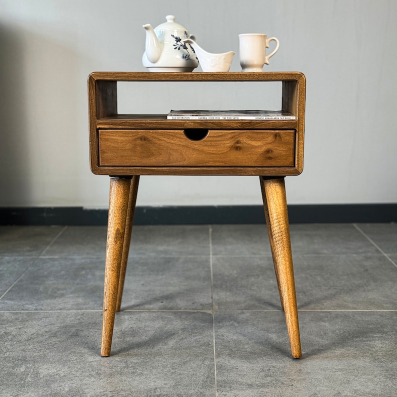 Op de afbeelding: Een houten bijzettafel met een lade en een plank. De tafel heeft een lichtbruine afwerking en taps toelopende poten. Een theepot, een kopje en een klein schaaltje staan op de plank.
