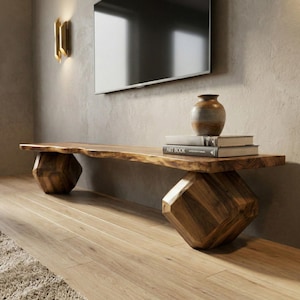 May include: A solid wood media console with a live edge top and geometric legs. The console is a natural wood color and holds a small vase and two books. The console is approximately 72 inches long.