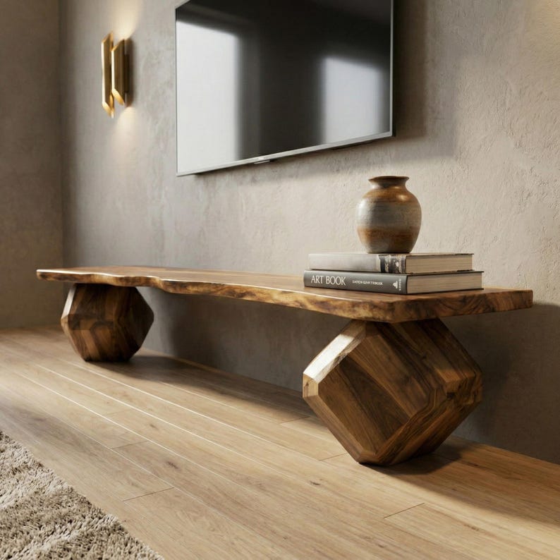 May include: A solid wood media console with a live edge top and geometric legs. The console is a natural wood color and holds a small vase and two books. The console is approximately 72 inches long.
