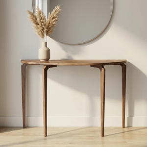 Peut inclure: Une console en bois de style moderne du milieu du siècle, avec un plateau en demi-lune et quatre pieds effilés. La table a une finition en bois naturel. Un vase beige avec des fleurs séchées est posé sur la table, avec un miroir rond en arrière-plan.
