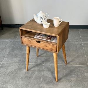 Op de afbeelding: Een houten bijzettafel met een lade en een plank. De tafel heeft een lichtbruine afwerking en is versierd met een witte theepot, een witte mok en een witte keramische lepel.