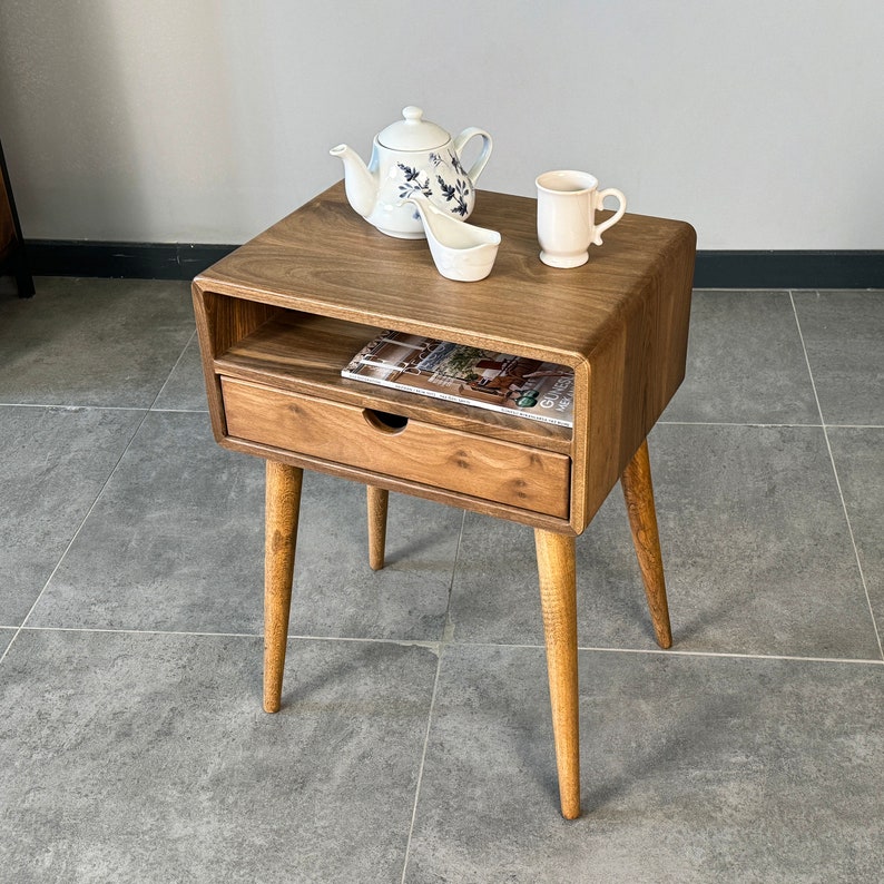 Op de afbeelding: Een houten bijzettafel met een lade en een plank. De tafel heeft een lichtbruine afwerking en is versierd met een witte theepot, een witte mok en een witte keramische lepel.