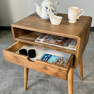 Op de afbeelding: Een houten bijzettafel met een lade en een plank. De tafel heeft een lichtbruine afwerking en afgeronde randen. De lade is open en bevat twee kleine zwarte containers en een tijdschrift. De plank is ook zichtbaar en bevat een tijdschrift. De tafel heeft vier poten die taps toelopen en lichtjes schuin staan.