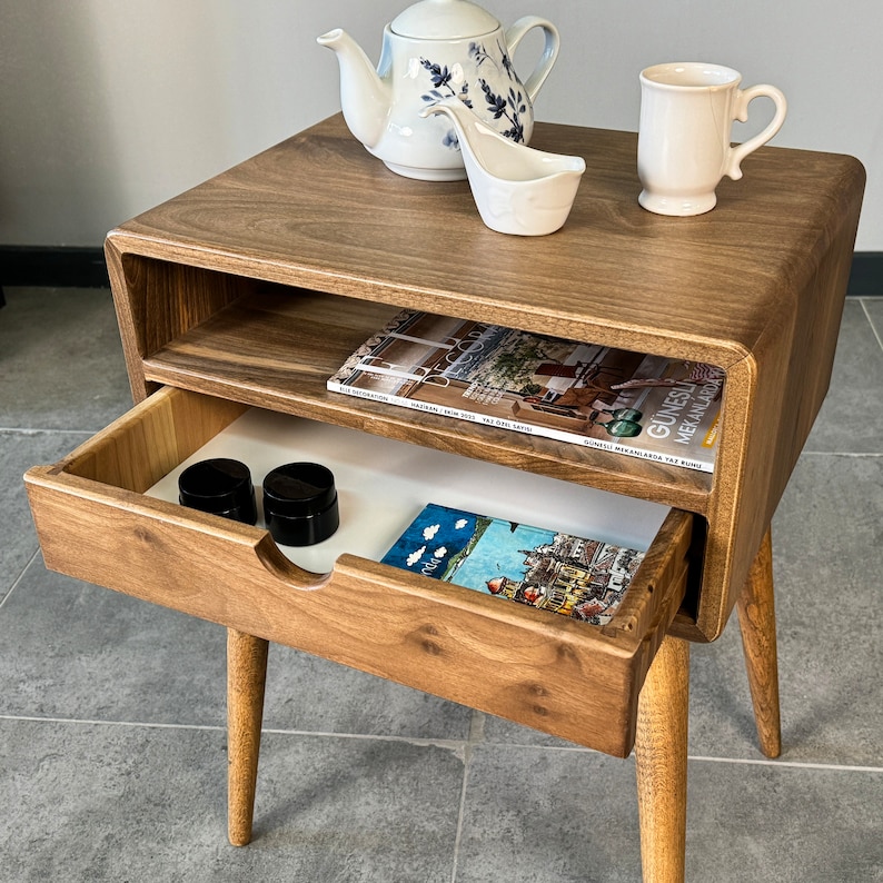 Op de afbeelding: Een houten bijzettafel met een lade en een plank. De tafel heeft een lichtbruine afwerking en afgeronde randen. De lade is open en bevat twee kleine zwarte containers en een tijdschrift. De plank is ook zichtbaar en bevat een tijdschrift. De tafel heeft vier poten die taps toelopen en lichtjes schuin staan.