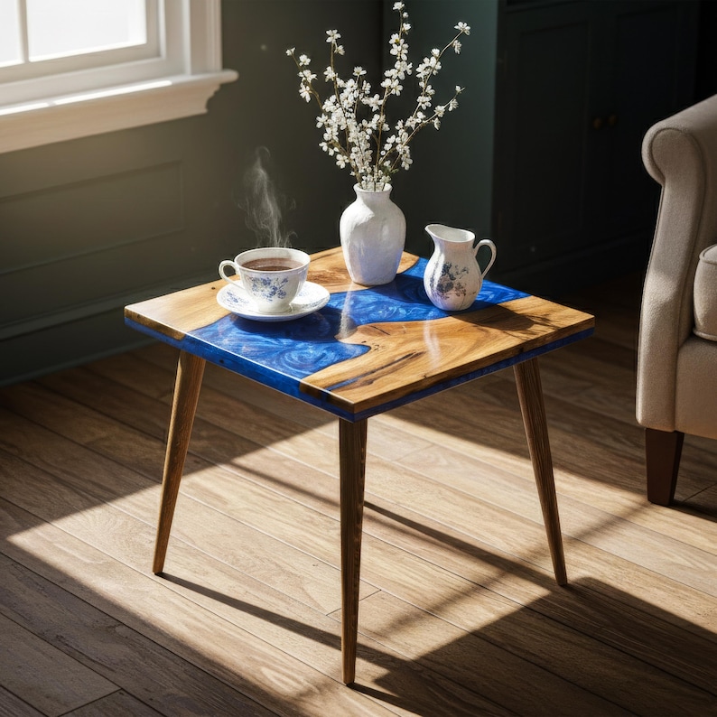 May include: A square wooden side table with a blue resin inlay. The table has four angled wooden legs. A white vase with flowers, a small creamer, and a teacup and saucer are on the table.