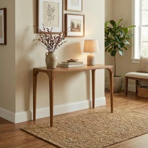 May include: A wooden console table with a curved design and four slender legs. A beige lamp sits on the table, along with books and a decorative vase with dried flowers. A woven rug is on the floor.