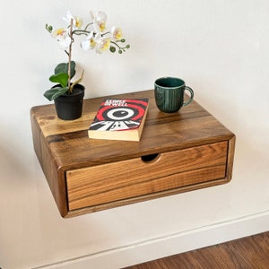May include: A wooden wall-mounted shelf with a drawer, featuring a book titled "George Orwell" and a green ceramic mug. A small potted plant with white flowers sits on the shelf.