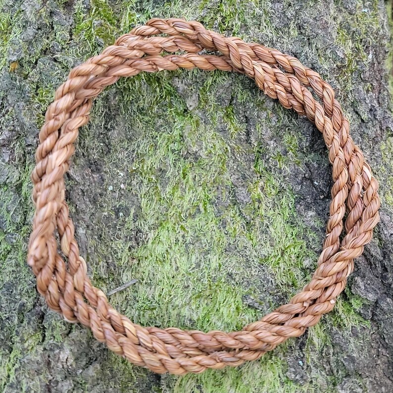May include: A circular, braided rope wreath made of natural fibers. The wreath is a warm brown color and is set against a backdrop of textured tree bark covered in green moss. The rope is tightly woven.