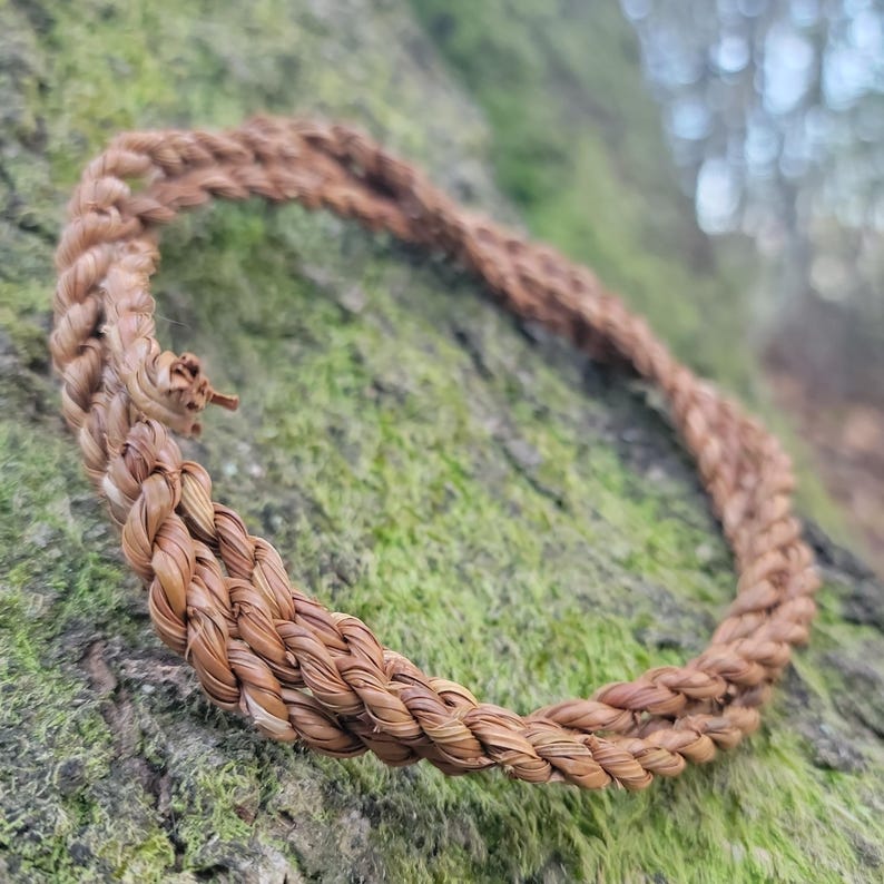 May include: A braided bracelet made of natural fibers, showcasing a warm brown hue. The bracelet is circular, with a textured surface, and rests on a mossy, textured background. The design suggests a rustic, handcrafted aesthetic.