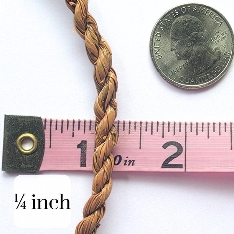 May include: Close-up of a braided, light brown rope, approximately 1/4 inch in diameter, next to a pink measuring tape and a US quarter. The rope is tightly woven and appears to be made of natural fibers. The measuring tape shows the rope's width.