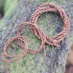 May include: Coiled brown rope made of natural fibers. The rope is arranged in a circular pattern with loose ends, suggesting it could be used for various crafts or decorative purposes. The rope rests on a moss-covered stone surface.