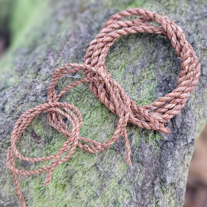 May include: Coiled brown rope made of natural fibers. The rope is arranged in a circular pattern with loose ends, suggesting it could be used for various crafts or decorative purposes. The rope rests on a moss-covered stone surface.