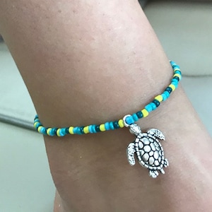 Op de afbeelding: Een blauwe en gele kralen enkelband met een zilveren schildpad bedel.