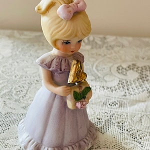 Puede incluir: Una figurita de porcelana de una ni&ntilde;a con un vestido lavanda que sostiene un n&uacute;mero cuatro dorado. Tiene el pelo rubio con un lazo rosa y mira hacia abajo al n&uacute;mero.