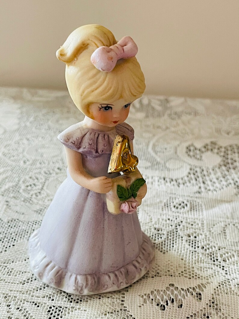 Puede incluir: Una figurita de porcelana de una ni&ntilde;a con un vestido lavanda que sostiene un n&uacute;mero cuatro dorado. Tiene el pelo rubio con un lazo rosa y mira hacia abajo al n&uacute;mero.